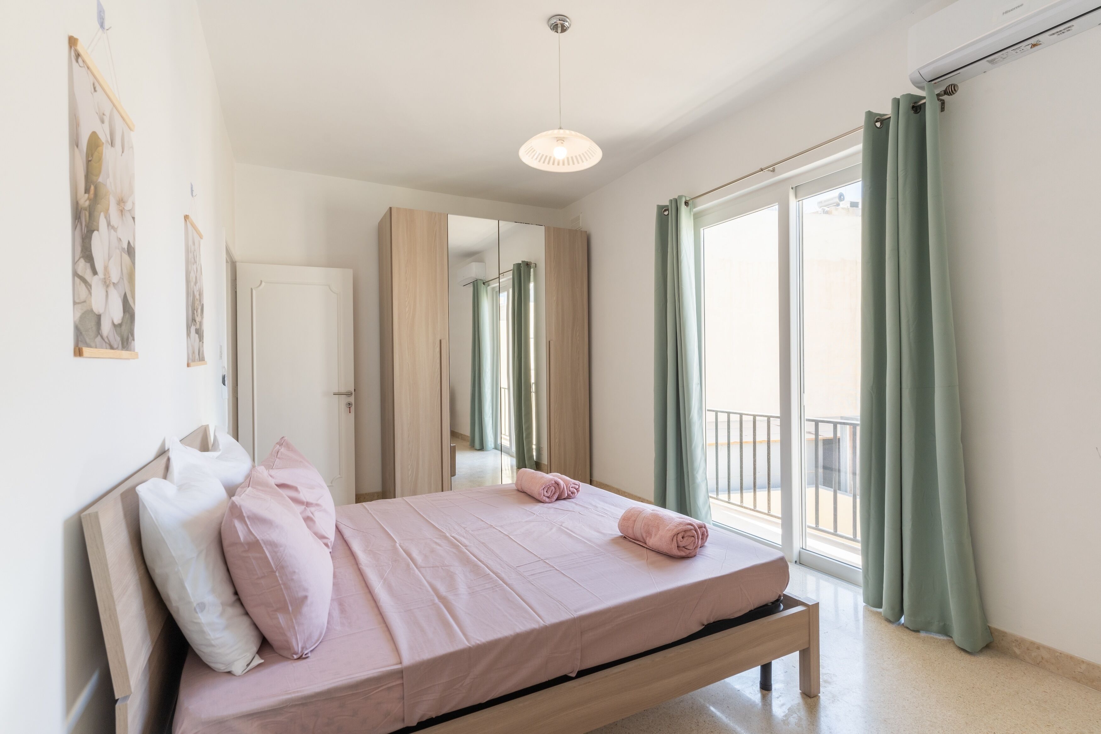 Un dormitorio con cama, almohadas, una mesita de noche, un espejo y vistas al exterior a través de una ventana con cortinas.