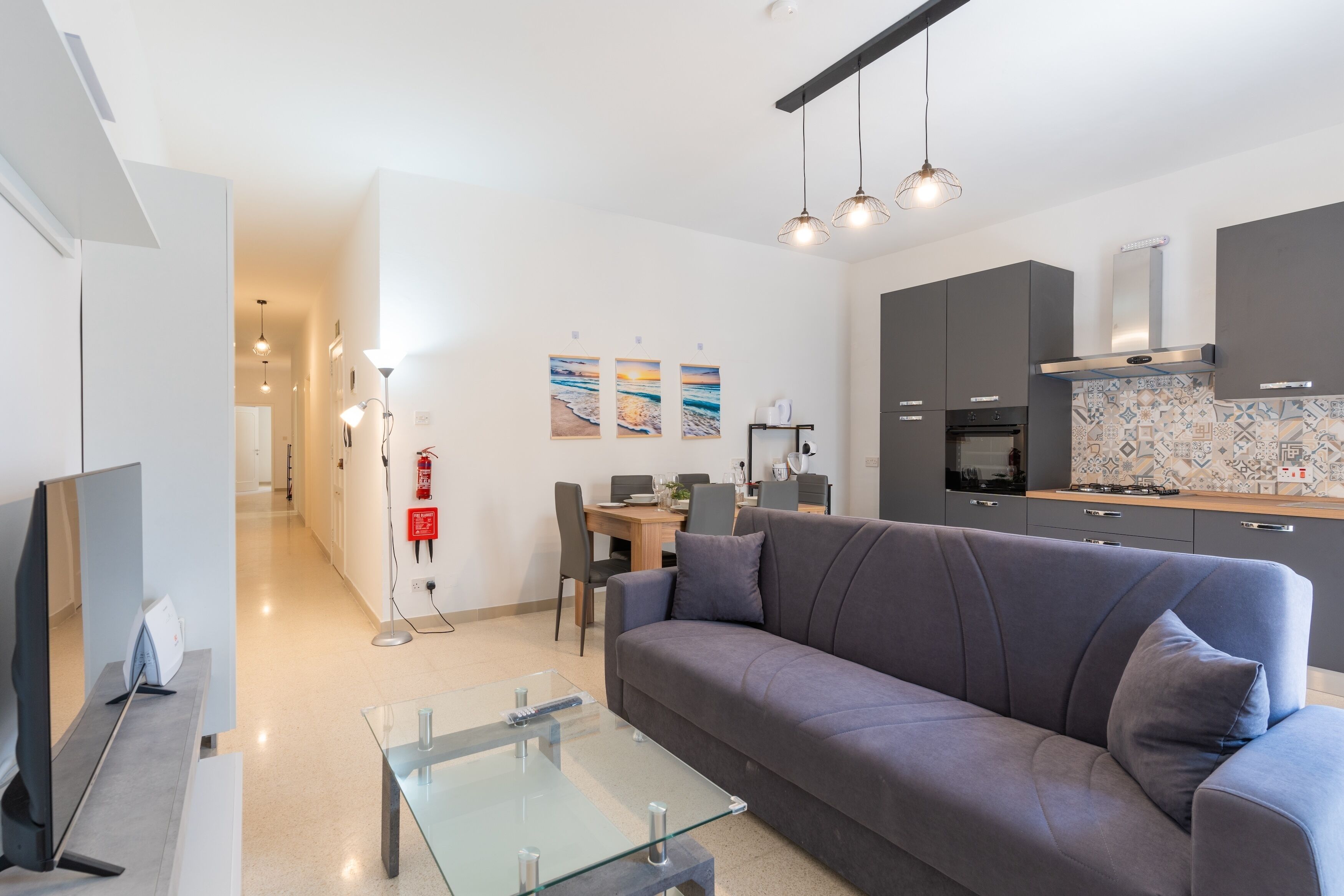 A modern living room with a grey sofa, a glass coffee table, and a TV on a stand. There is a kitchen area with dark cabinets and a tiled backsplash.