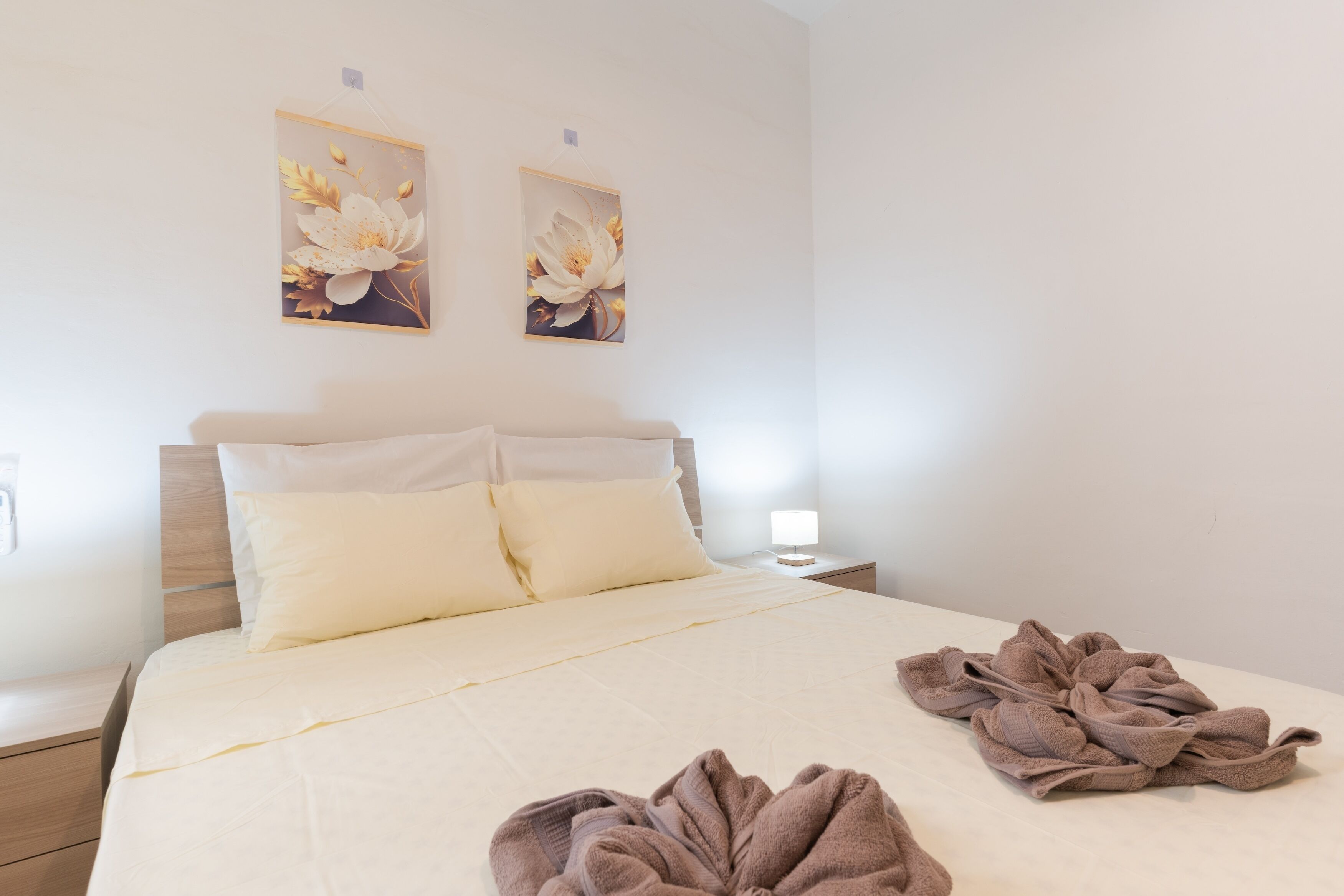 Een netjes opgemaakt bed met witte beddengoed en twee kussens, een nachtkastje met een lamp, en twee bloemrijke wanddecoraties.