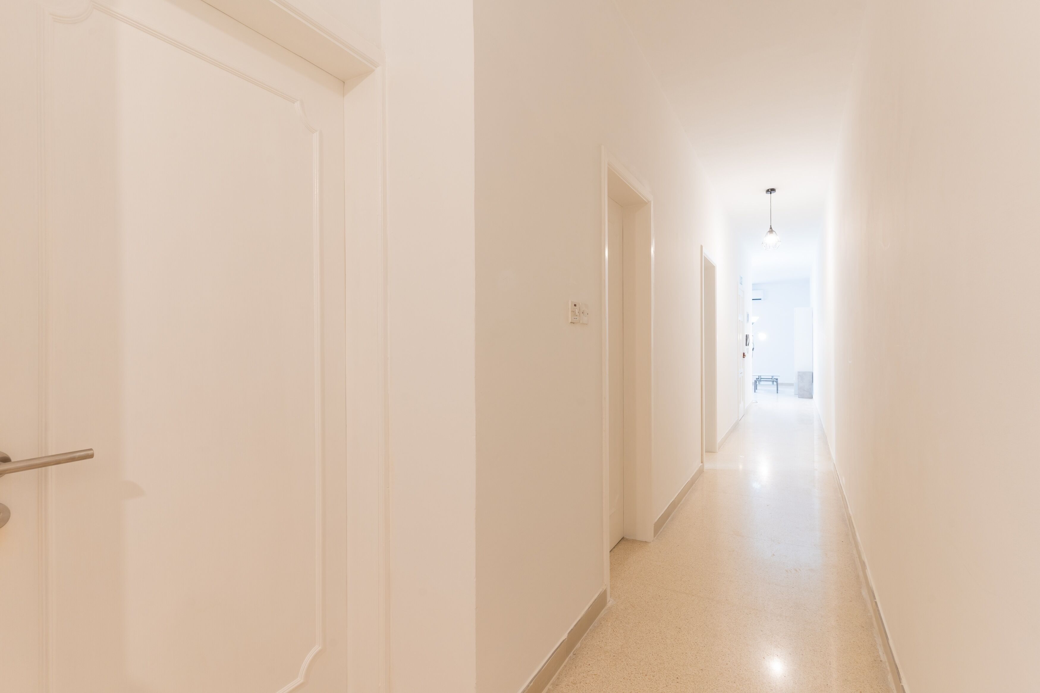 Un couloir étroit et bien éclairé, aux murs blancs et au sol de couleur claire, qui mène à une pièce lumineuse au bout.