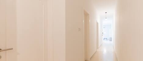 Un pasillo estrecho y bien iluminado con paredes blancas y suelo de tono claro, que conduce a una habitación luminosa al final.