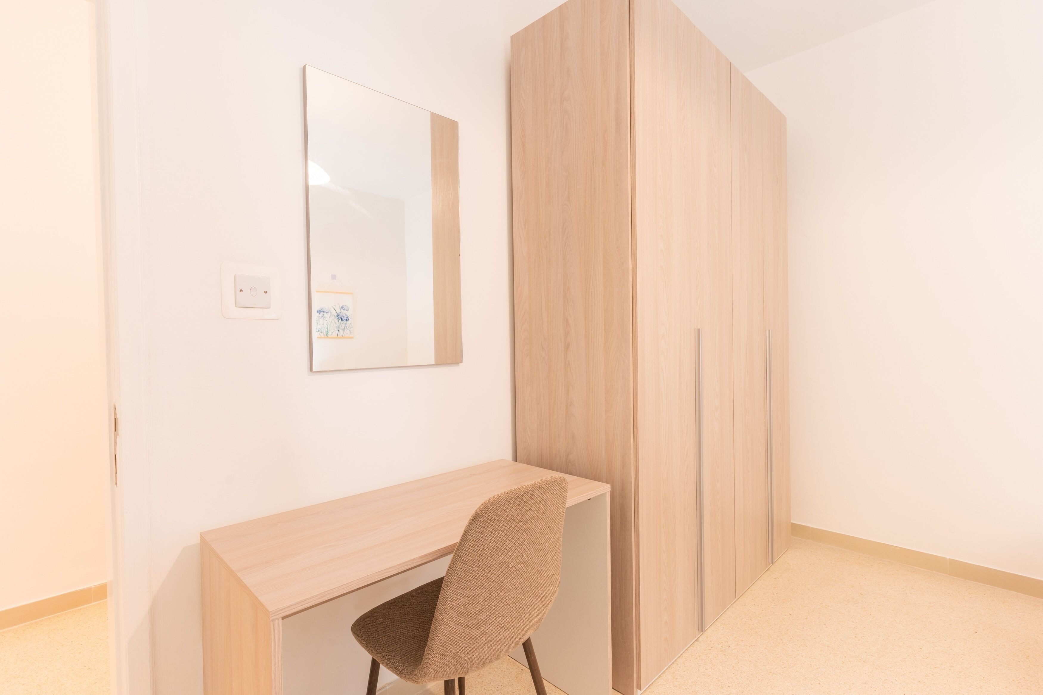 A room with a wooden desk, a chair, a wardrobe, and a mirror on the wall.