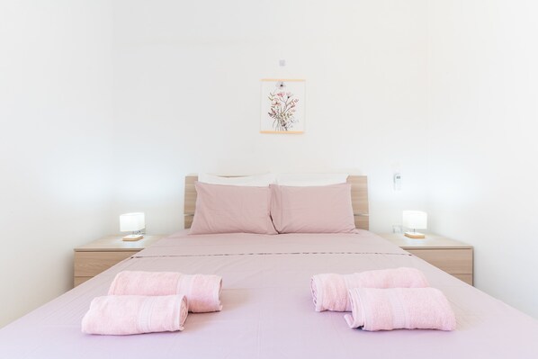 Un lit bien fait, avec une literie et des serviettes roses, une œuvre d’art florale encadrée au mur et des lampes de chevet posées sur des tables de nuit en bois.