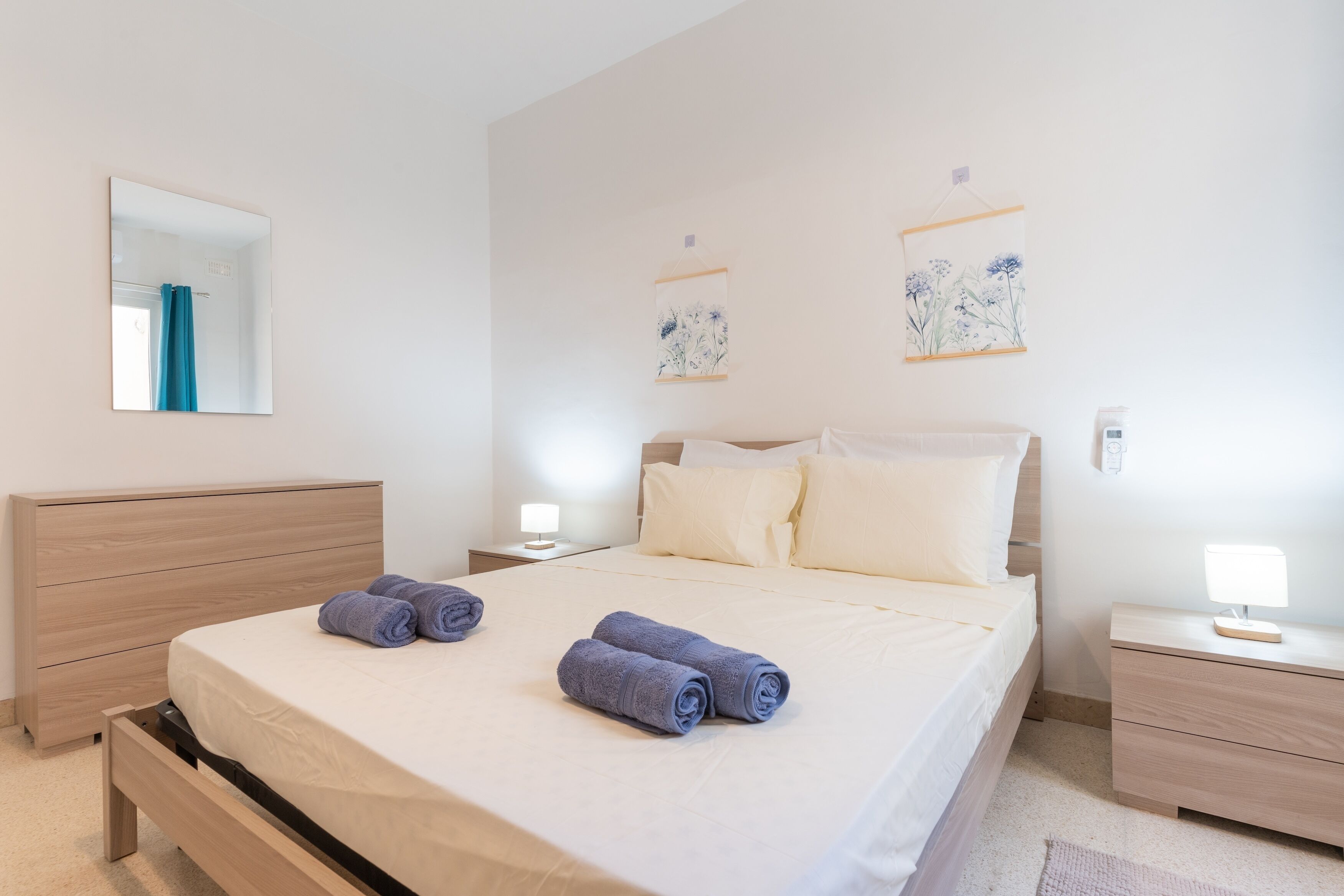Una cama bien hecha con dos toallas enrolladas, una mesita de noche con lámpara, un taburete, un aparador de madera y un espejo instalado en la pared.