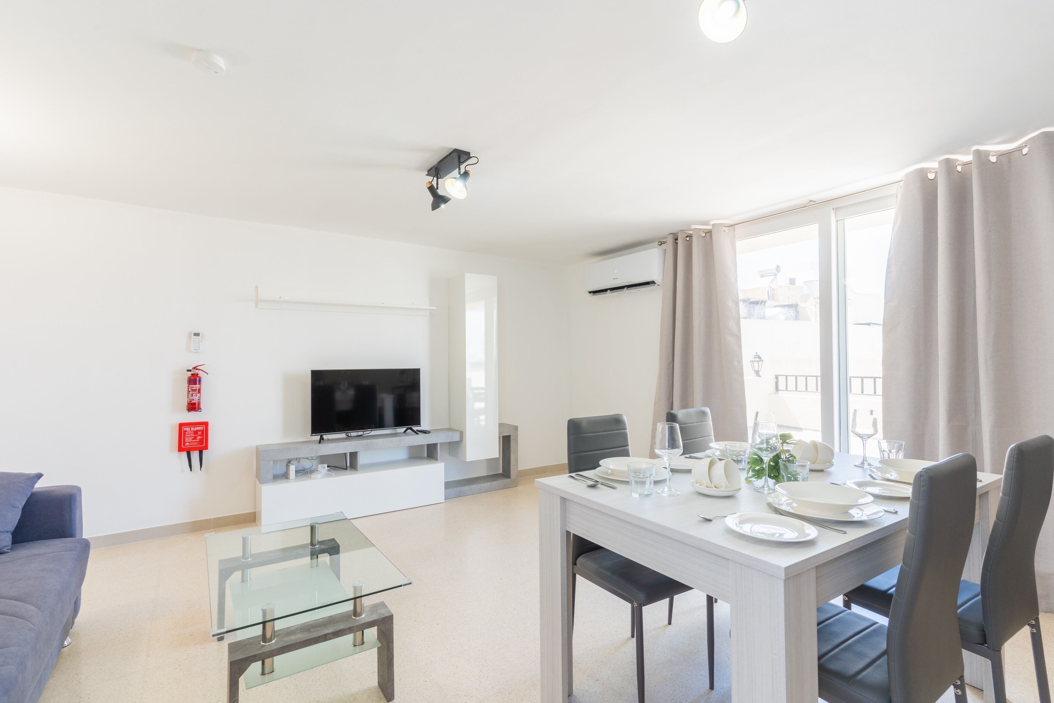 Een moderne woonkamer met een flatscreen televisie, een glazen salontafel, een bank en een eethoek compleet met borden, glazen en een plant.
