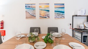 A dining area with a wooden table set for four, three framed ocean sunset paintings on the wall, and a fire extinguisher on the left. - Tempo Suites (St. Paul's Bay)