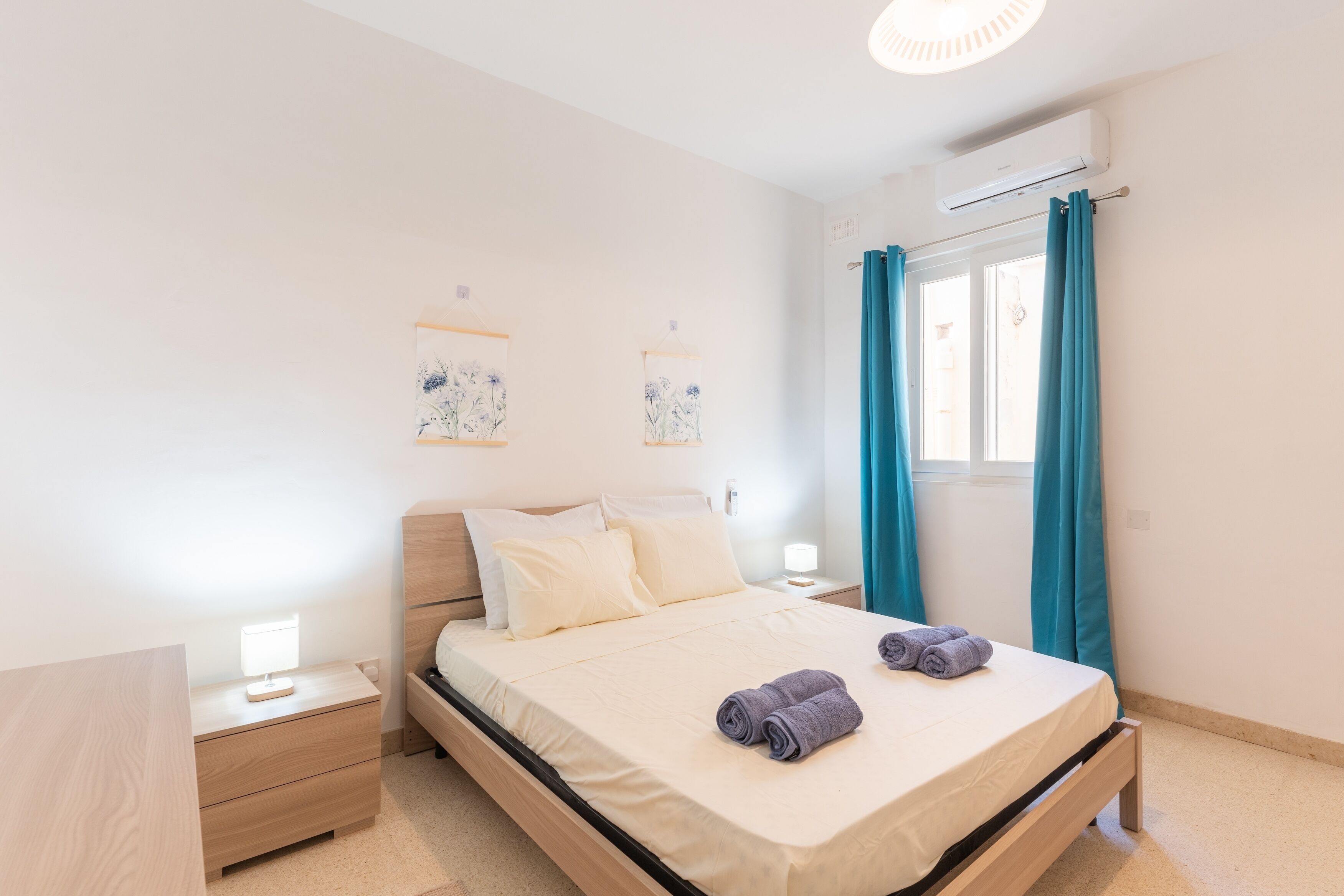 Ein Schlafzimmer mit Bett, Nachttischen, einer Fensterbank, Vorhängen und Wandbildern.