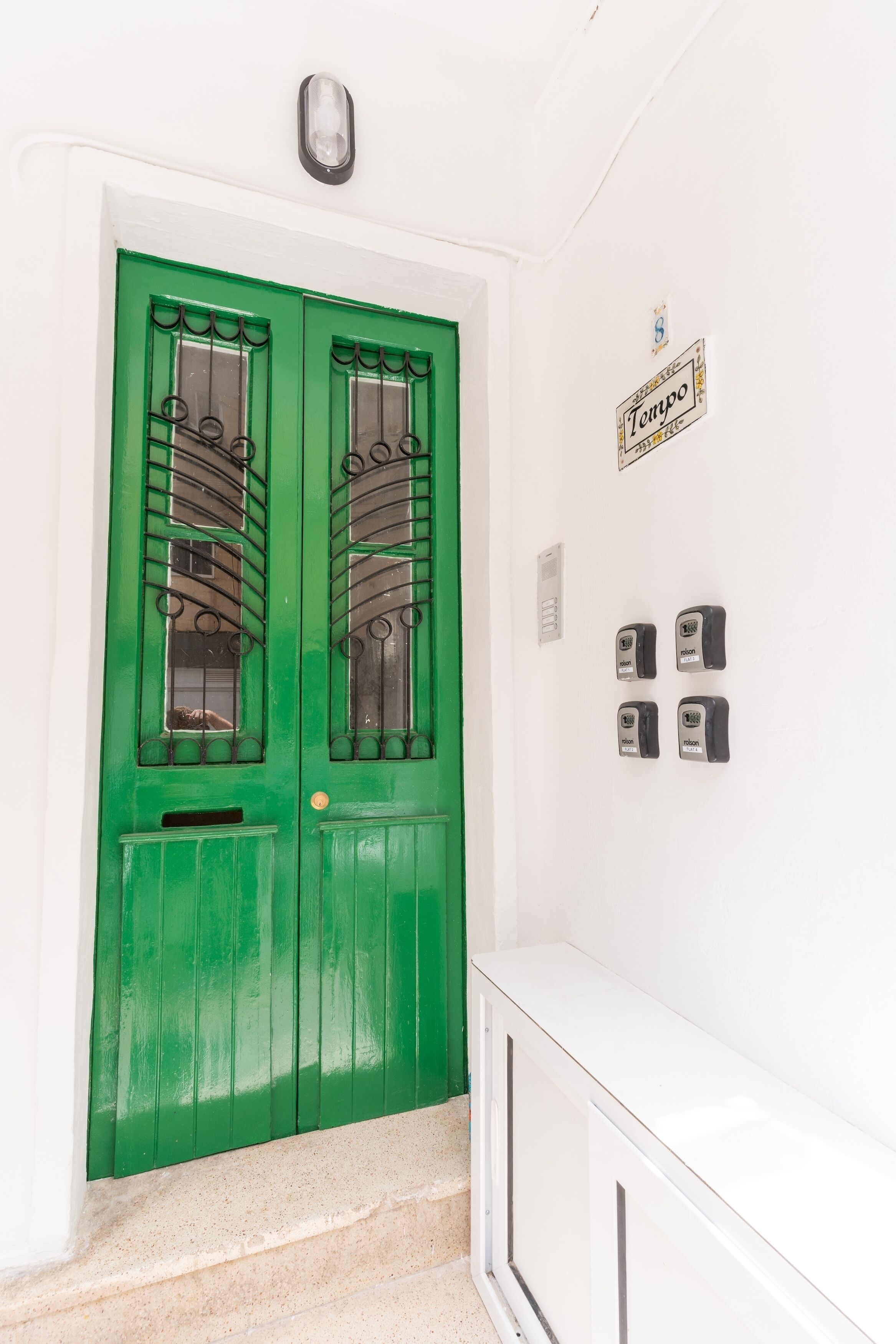 Une porte verte ornée d’une grille métallique, un mur blanc sur lequel est apposée une enseigne « Tempo », et une console blanche.