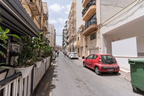 A narrow urban street lined with multi-story buildings, parked cars, and potted plants along the sidewalk. - Tempo Suites (St. Paul's Bay)