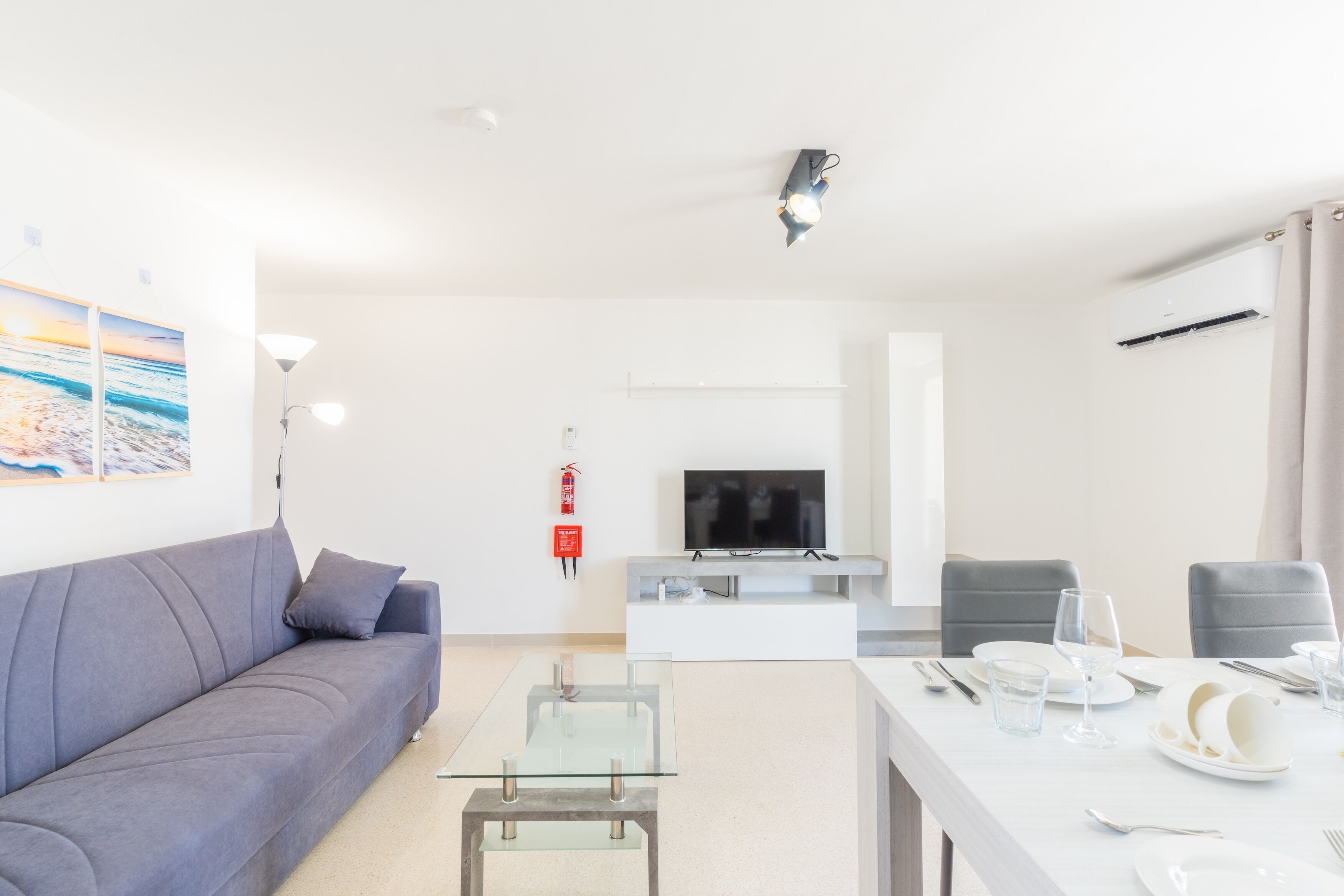 Un salon moderne avec un canapé gris, une table basse en verre, un téléviseur à écran plat et un coin repas avec une table blanche mise pour deux personnes.