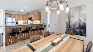 A modern kitchen with wooden cabinets, a stainless steel refrigerator, and a bar area. There is a dining table with chairs and a chandelier above.