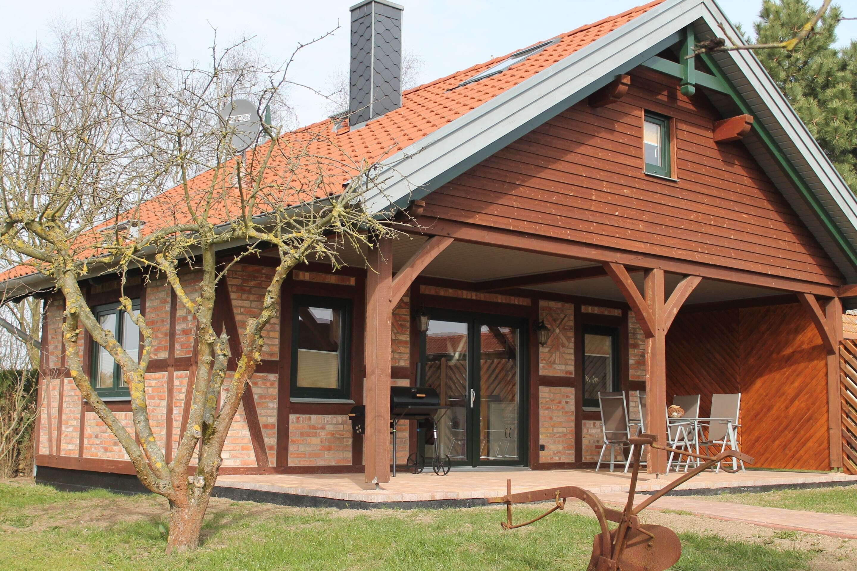 Une maison en bois avec un toit en pente, une cheminée en briques et une véranda couverte avec des sièges d’extérieur.