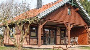 A wooden house with a sloped roof, a brick chimney, and a covered porch with outdoor seating. - Modern Holiday Home in Brusow With Roofed Terrace (Kroepelin)