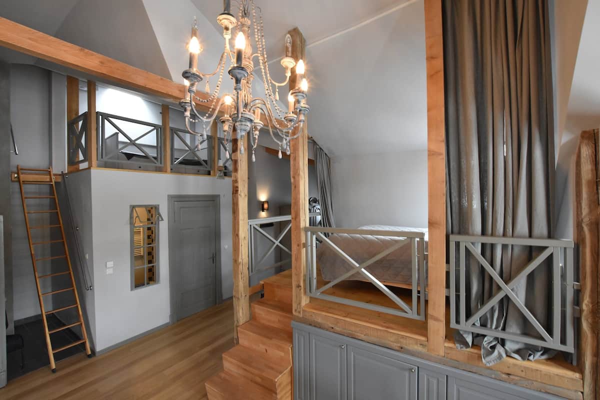 Ein Schlafzimmer im Loft-Stil mit freiliegenden Holzbalken, einer Kristallleuchtre und einem Schlafbett auf Podest.