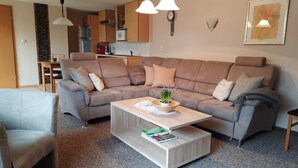Ein modernes Wohnzimmer mit einem Ecksofa, einem Couchtisch mit Zeitschriften und einer Küche im Hintergrund.
