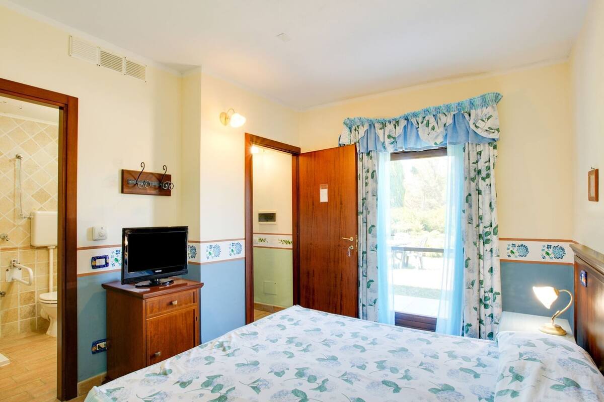 Une chambre à coucher avec un lit, un téléviseur posé sur une commode en bois, une salle de bain avec douche et un balcon avec des rideaux.