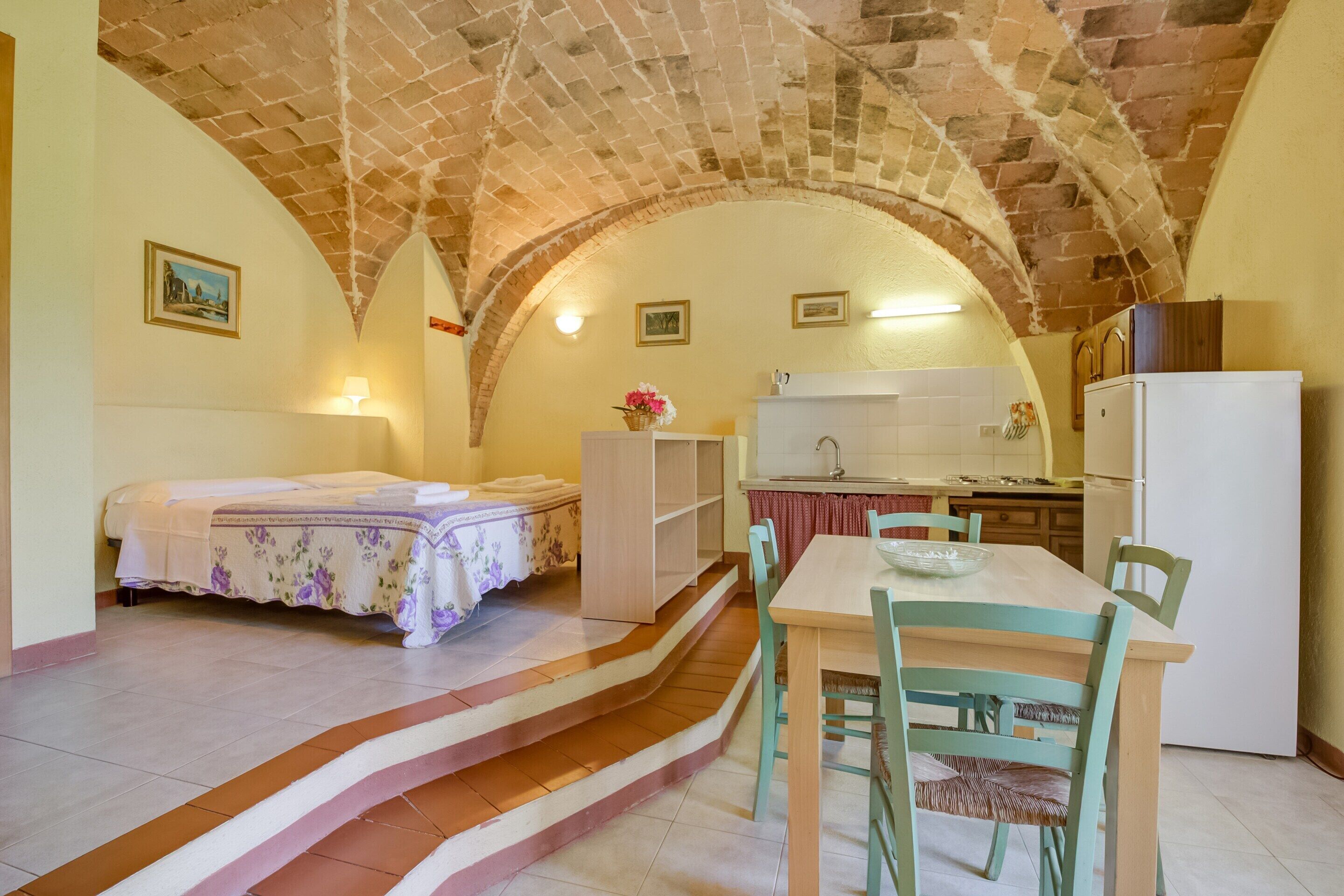 A room with a bed, a dining table with chairs, a kitchen area, and a staircase leading to an upper level.