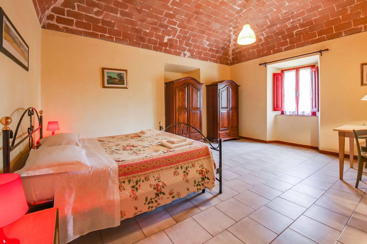 Een slaapkamer met een bakstenen plafond, een bed met een bloemenprintdeken, een houten garderobe, een rode lamp en een raam met rode gordijnen.