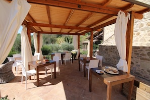 An outdoor patio with wooden tables and white chairs, surrounded by stone walls and greenery.