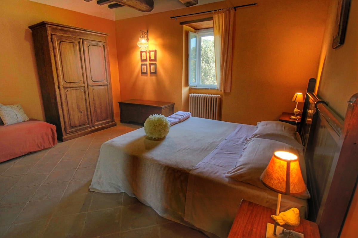 Un dormitorio con cama, mesita de noche, lámpara, ventana con cortinas, radiador y armario.