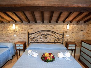 A bedroom with a stone wall, a wooden ceiling, a metal headboard, two bedside tables, a bed with blue bedding, and a bouquet of flowers on the bed. - Holiday Home in Castiglione del With Pool (Castiglione del Lago)