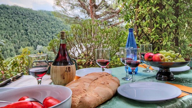 An outdoor dining setup with a table set for a meal, including bread, a bowl of tomatoes, and a bottle of red wine. - Country House With Swimming Pool and Garden With Mediterranean Plants (Bettona)