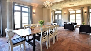Un vaste espace dédié à la salle à manger, avec une grande table en bois, des chaises blanches et un lustre. On aperçoit deux canapés noirs au fond de la pièce, et de grandes fenêtres, ornées de rideaux, illuminent l’ensemble.