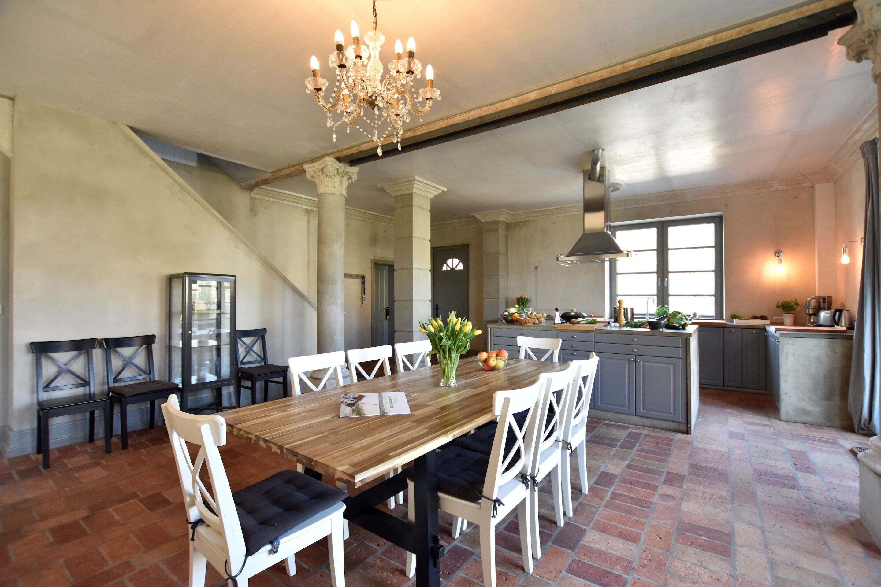 Een ruime eethoek met een houten tafel en witte stoelen, een kroonluchter en een keuken op de achtergrond.