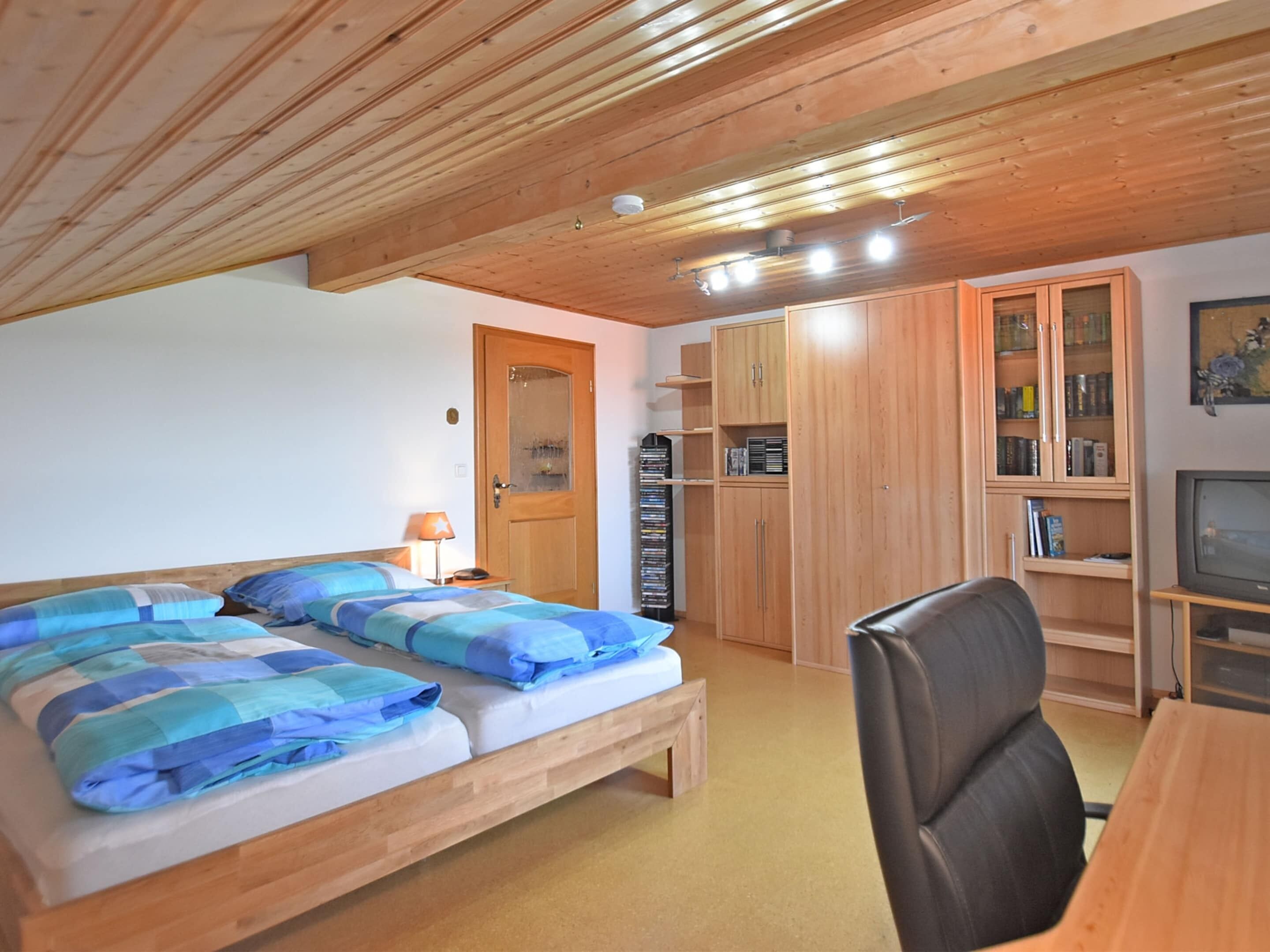 Una habitación con dos camas, techo de madera, un escritorio, una silla, un televisor y una estantería.