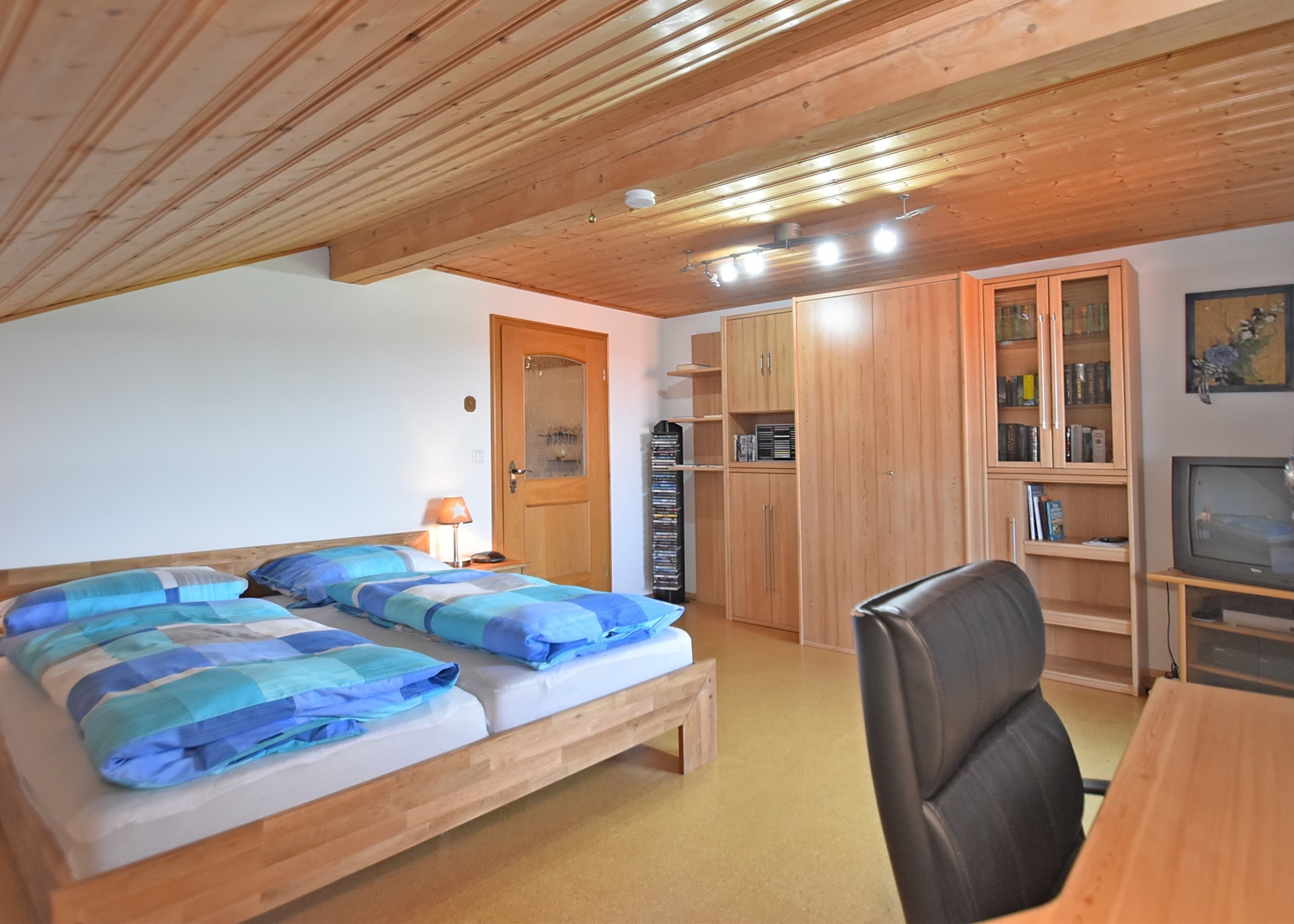 Ein Schlafzimmer mit Holzbalkendecke, einem Bett mit blauer Bettwäsche, einem Schreibtisch mit Stuhl und einem Fernseher.