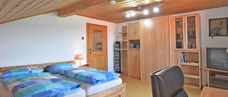 A bedroom with a wooden ceiling, a bed with blue bedding, a desk with a chair, and a TV.