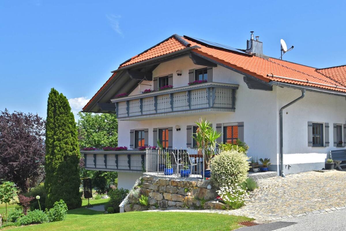 Une maison à deux étages, avec un toit recouvert de tuiles rouges, un balcon doté de balustrades et un escalier en pierre menant à l’entrée.