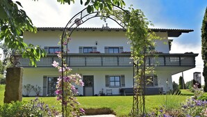 Ein zweigeschossiges Haus mit Balkon, umgeben von einem Garten mit verschiedenen Pflanzen und Blumen.