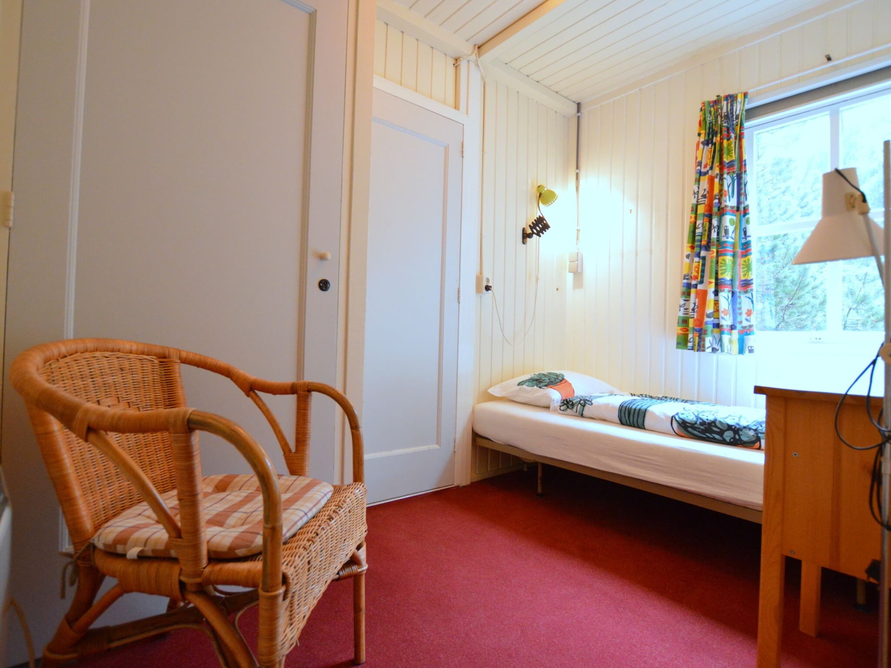 A small room with a single bed, a wicker chair, a door, and a window with curtains.
