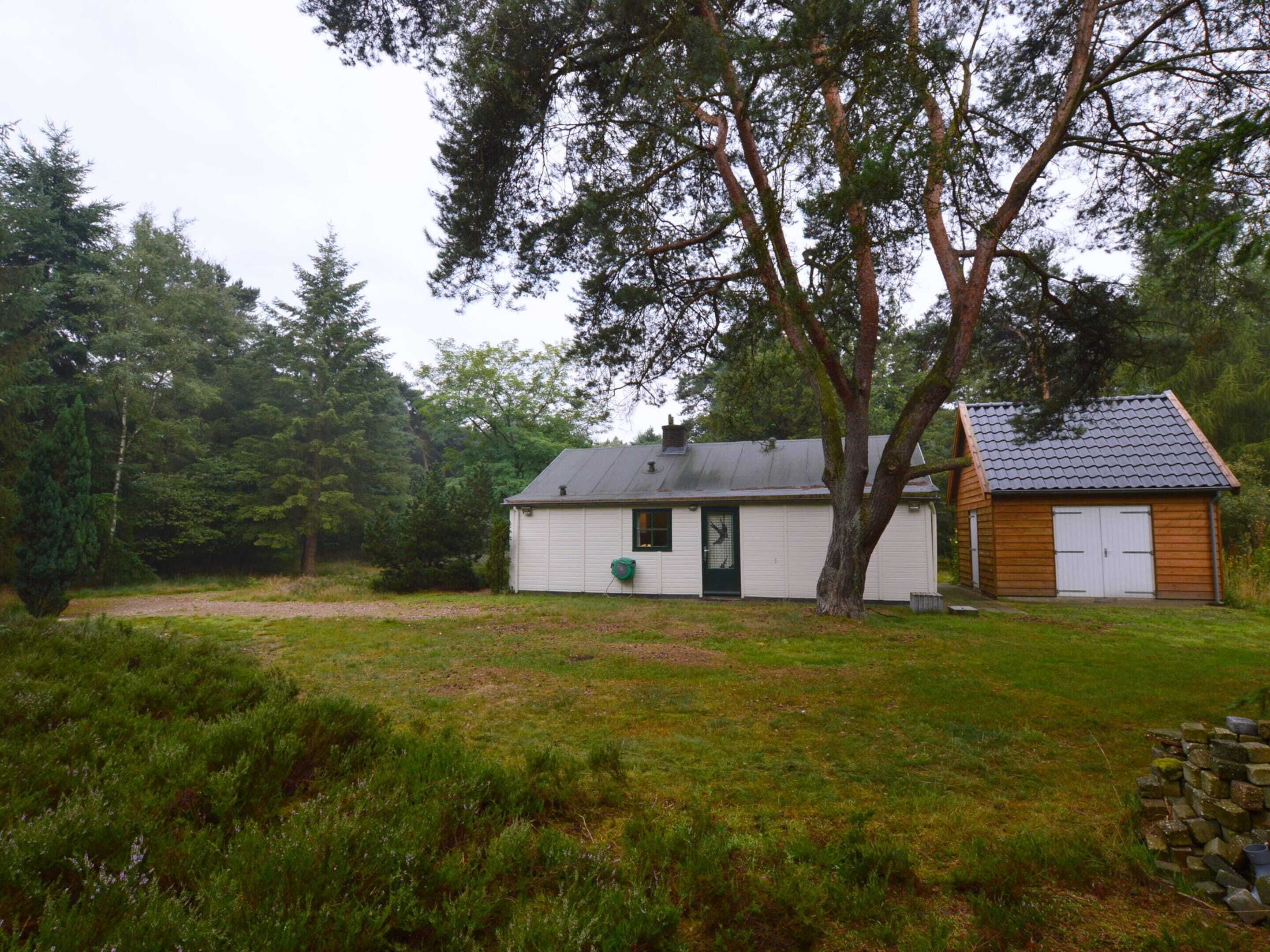 A small, single-story house with a garage, surrounded by a grassy yard and dense trees.