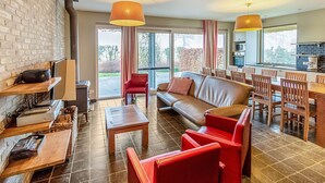 A modern living room with a sofa, chairs, a coffee table, and a dining area with a table and chairs. There is a fireplace, a large window, and a kitchen area visible through the glass doors. - Ideal Holiday Home in Gijverinkhove With Terrace, Garden (Alveringem)