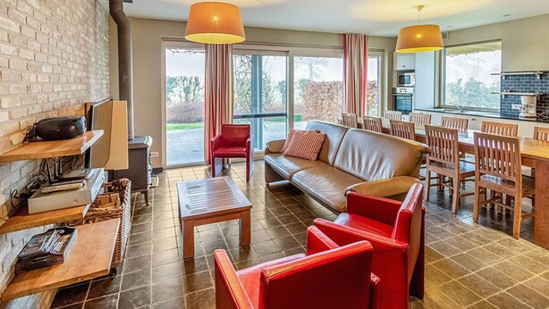 Un salon moderne comprenant un canapé, des fauteuils, une table basse et un coin repas avec une table et des chaises. On y trouve également une cheminée, une grande fenêtre et une cuisine visible à travers les portes vitrées.
