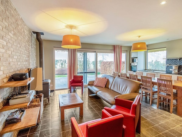 Un salon moderne comprenant un canapé, des fauteuils, une table basse et un coin repas avec une table et des chaises. On y trouve également une cheminée, une grande fenêtre et une cuisine visible à travers les portes vitrées.
