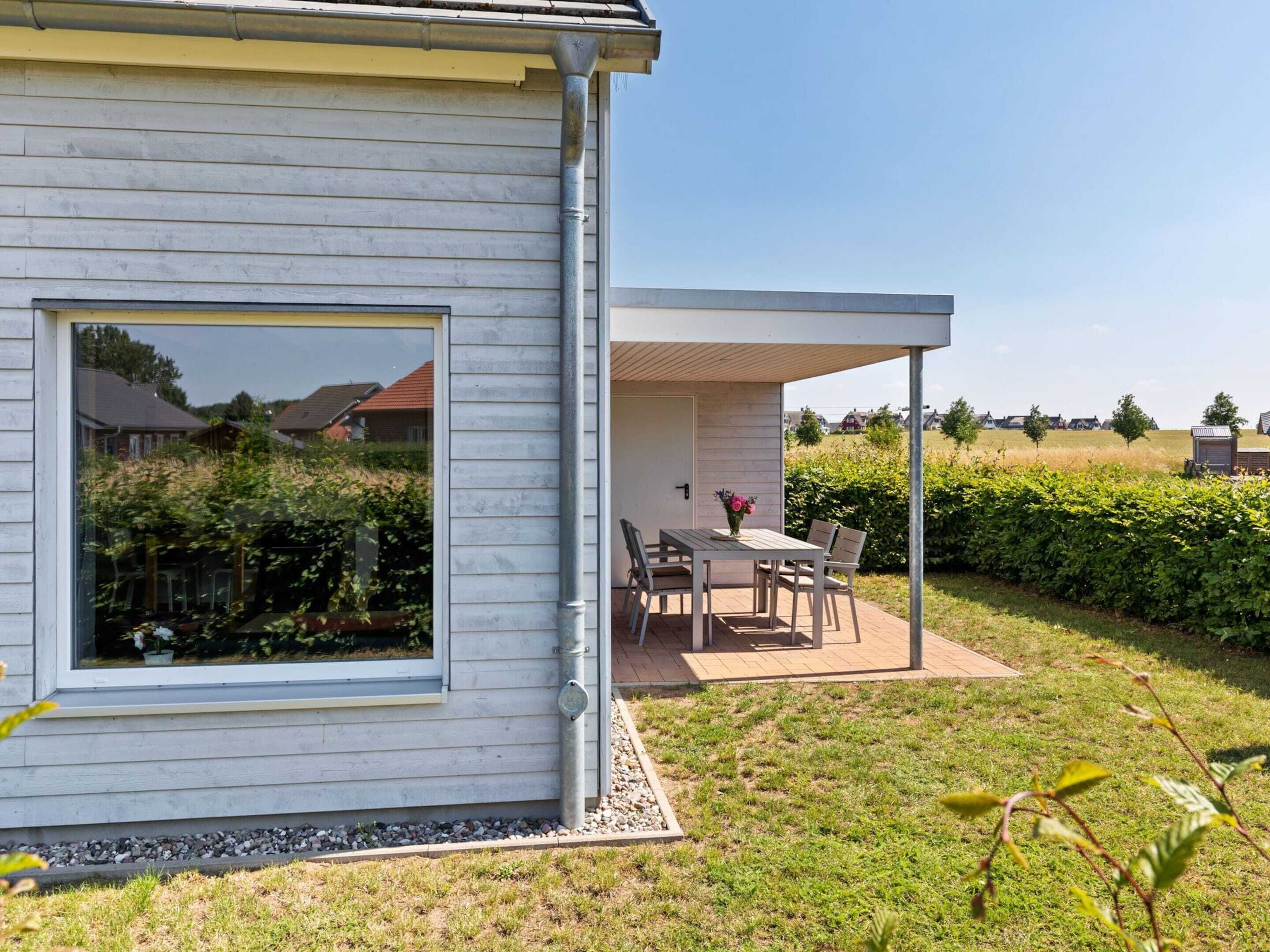 A modern house with a large window, a covered patio with a table and chairs, and a view of the surrounding landscape.