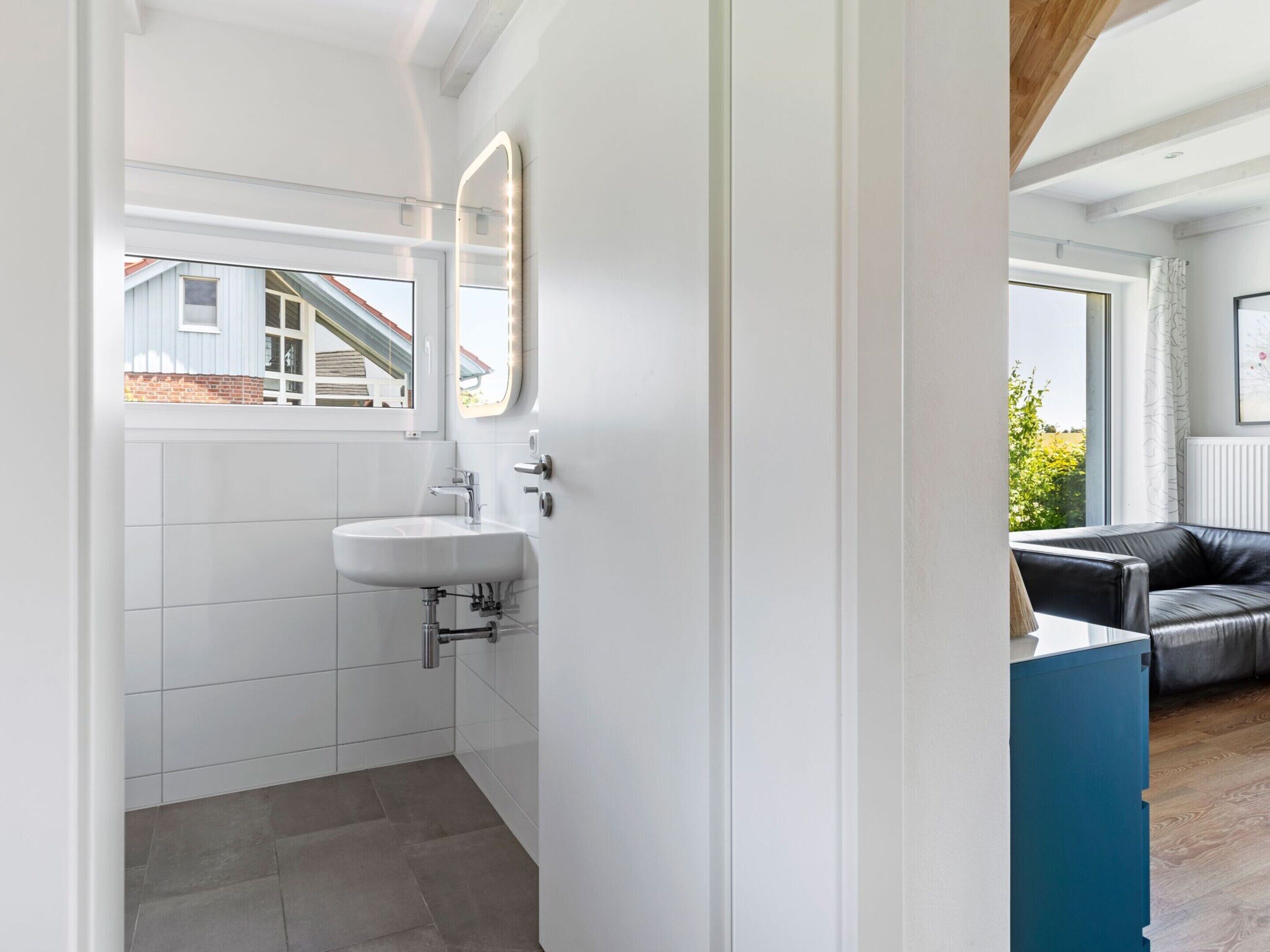 A modern bathroom with a white sink, a mirror with lights, and a view of a house through a window.