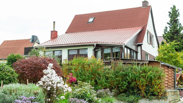 Una casa de dos pisos con techo de tejas rojas, rodeada de plantas y flores diversas.