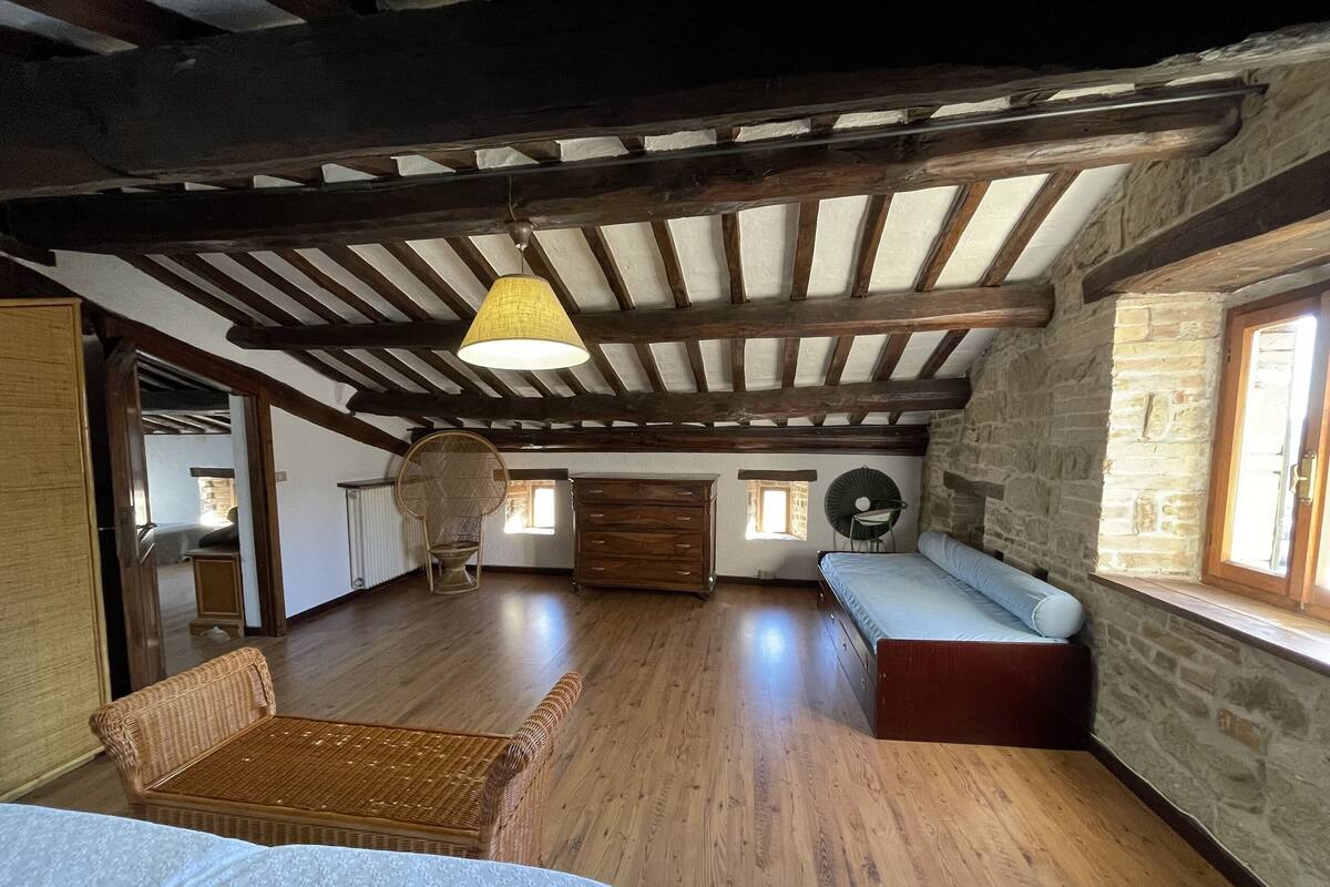 A spacious attic room with exposed wooden beams, a large window, a wicker chair, a wooden dresser, and a bed with a white bedspread.