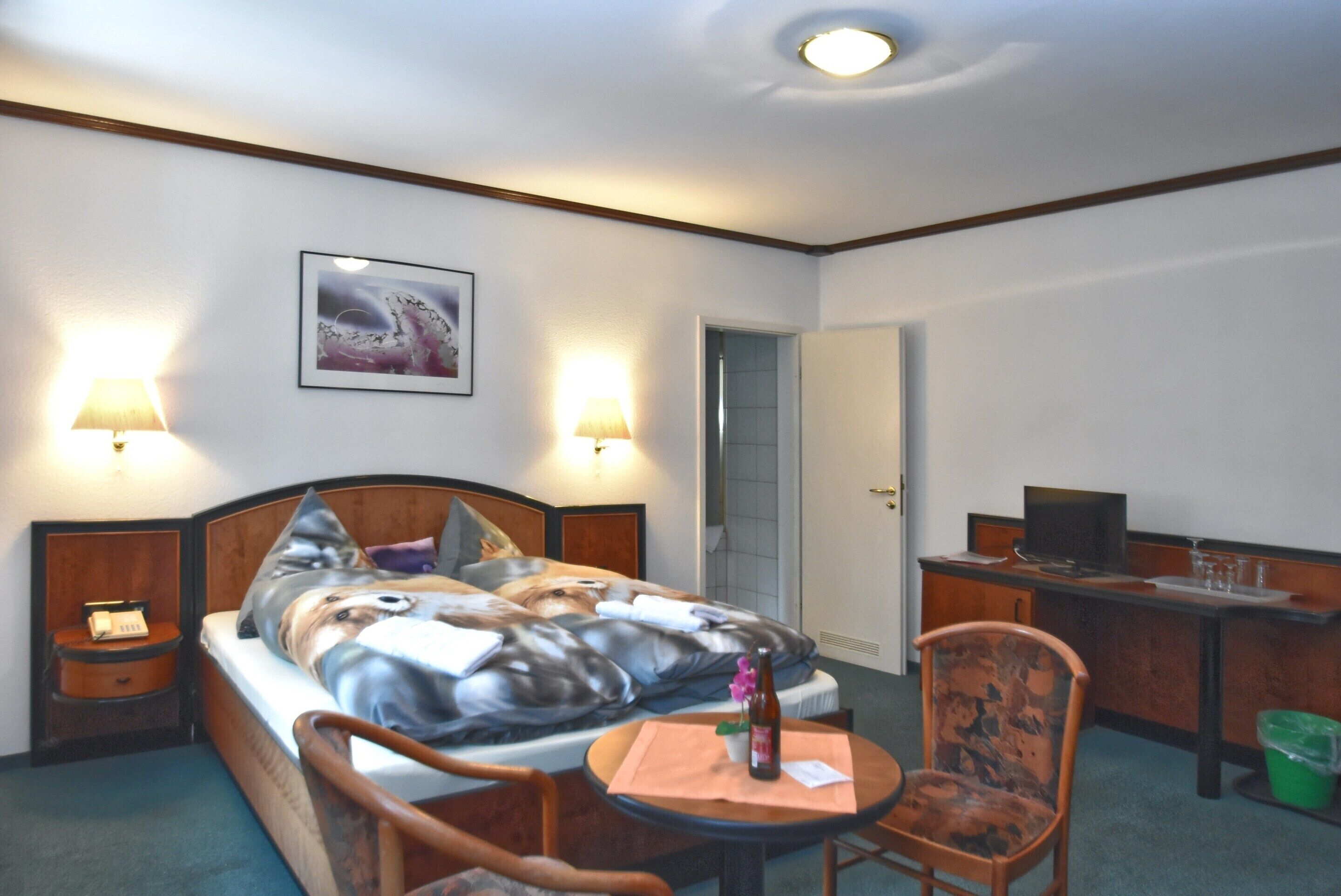 A hotel room with a bed, bedside lamps, a desk with a computer, a chair, and a painting on the wall.
