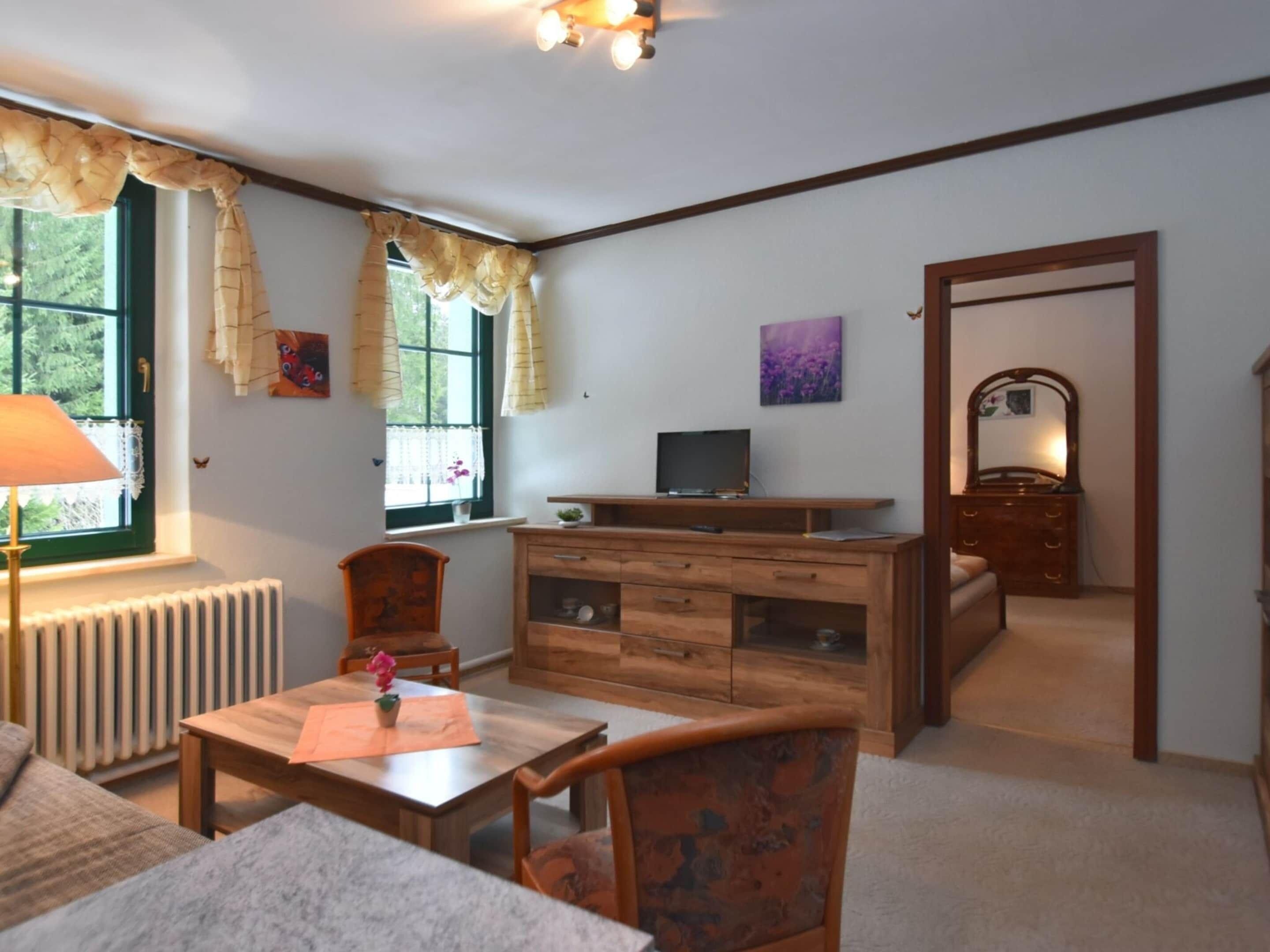 A cozy room with a wooden dresser, a television on a stand, a chair, a table with a vase, and a window with curtains.