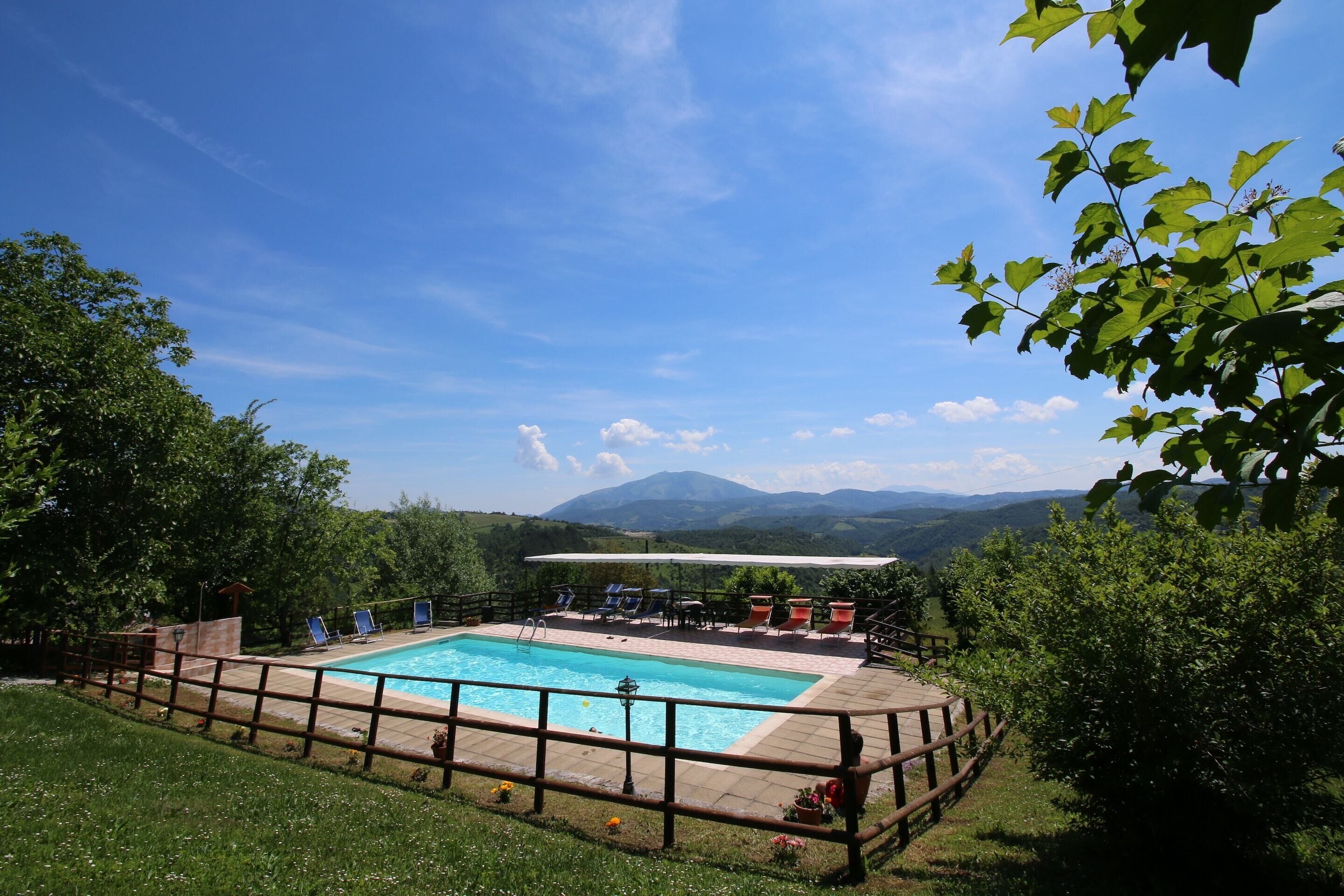 Ein Pool mit Holzterrasse und Liegestühlen, umgeben von Grünflächen und Bergen im Hintergrund.