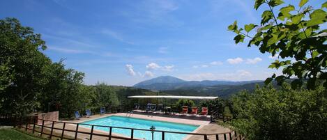 Ein Pool mit Holzterrasse und Liegestühlen, umgeben von Grünflächen und Bergen im Hintergrund.