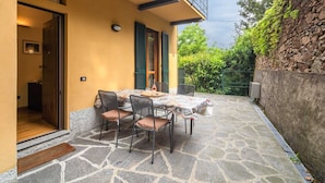 A patio with a table and chairs, a door leading to a room, and a stone wall with greenery. - Modern Apartment in Meina Near the Lake (Meina)