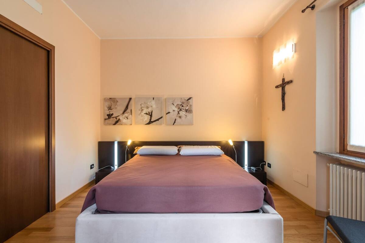 A bedroom with a bed, wooden floor, wall-mounted artwork, a cross, and a window with a radiator.