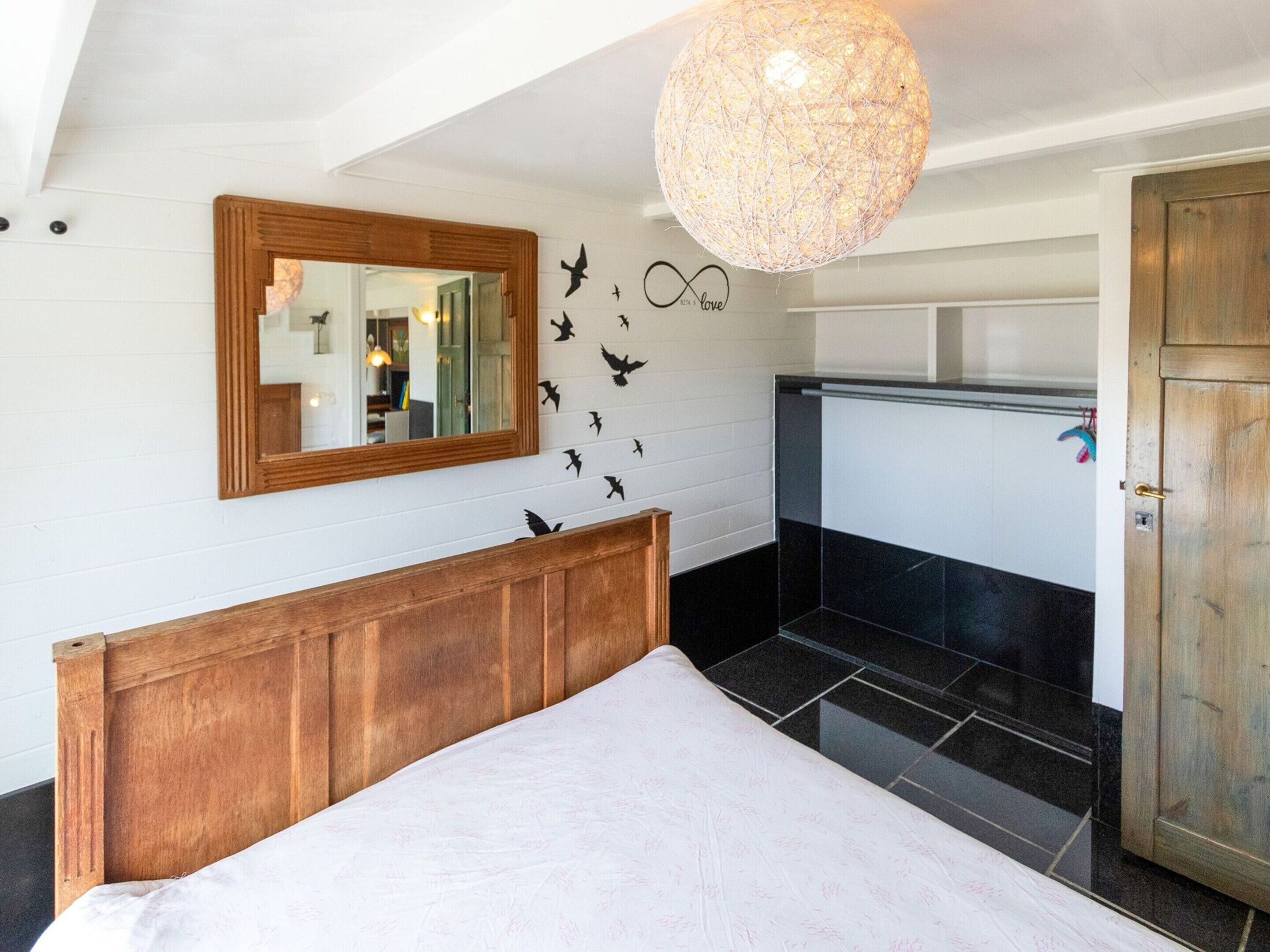 A bedroom with a wooden headboard, a large mirror, a wall-mounted bird decal, a hanging light fixture, and a door with a handle.