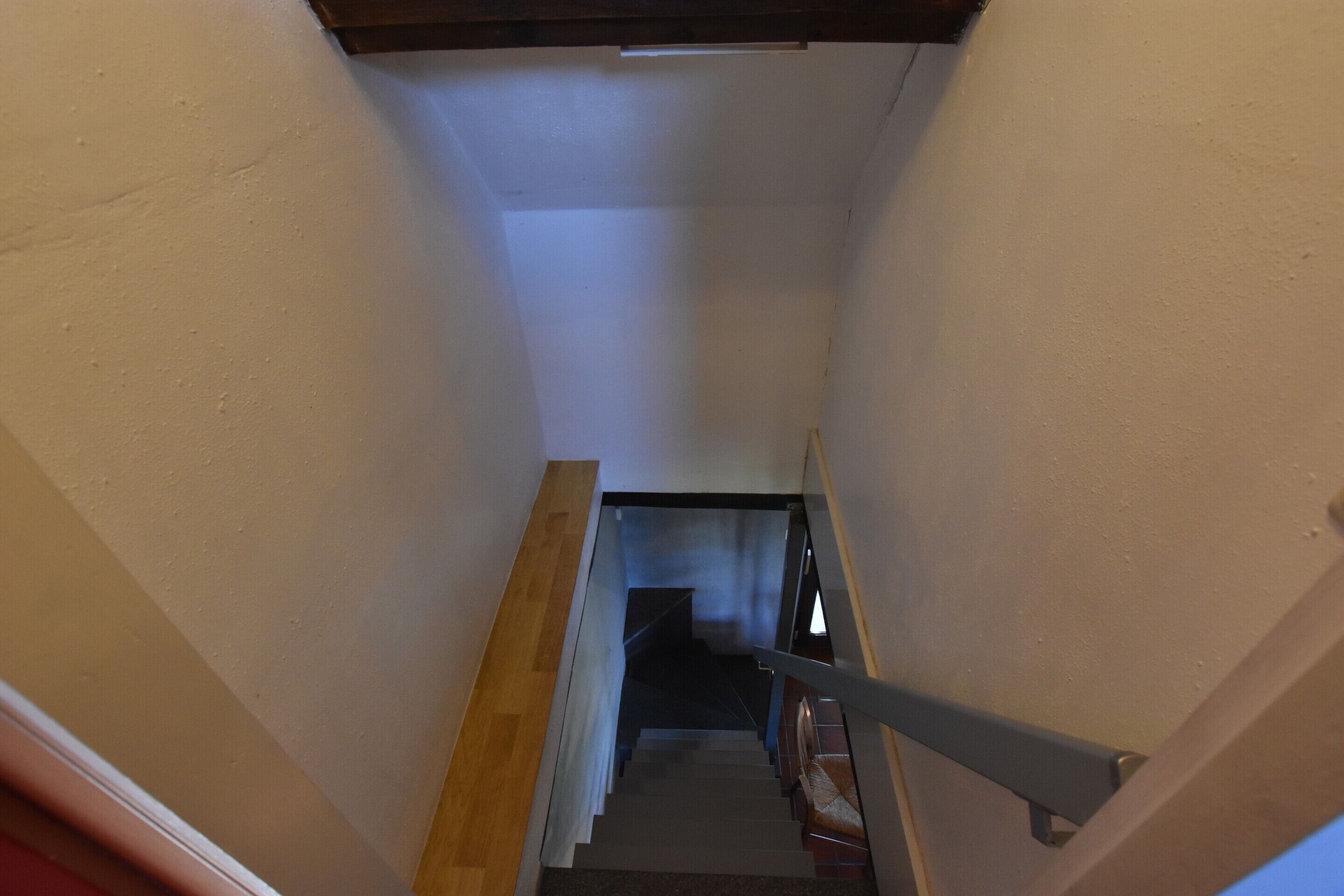 A narrow stairway with a wooden handrail and a metal railing, leading to a landing with a dark-colored staircase.